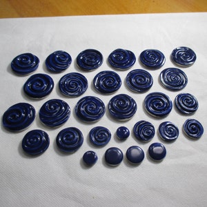 Op de afbeelding: Een verzameling van 25 blauwe keramische knopen met een swirl-ontwerp. De knopen zijn in een halve cirkel op een wit oppervlak gerangschikt.