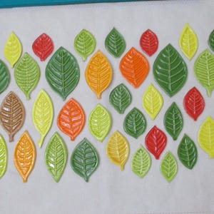 May include: A collection of colorful ceramic leaf decorations in various sizes and shades of green, yellow, orange, red, and brown. The leaves are arranged on a white surface, creating a decorative pattern. Ideal for home decor or craft projects.