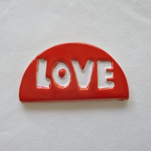 May include: A red, semi-circular decorative object with the word "LOVE" in white, block letters. The object has a glossy finish and is set against a white, textured background. The letters are cut out, revealing the white background.
