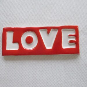 May include: A rectangular, red ceramic tile with the word "LOVE" in white, block letters. The letters are raised and the tile has a glossy finish. The tile is a decorative item.