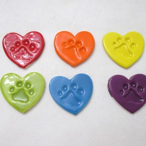 May include: Six heart-shaped ornaments in red, orange, yellow, light green, blue, and purple. Each heart features a debossed paw print impression on its smooth, glossy surface. These decorative items are arranged in two rows of three.