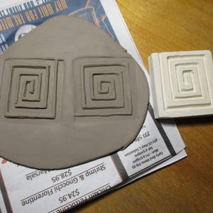 May include: A light brown clay piece with two square spiral designs and a white square stamp with a spiral design. The clay piece is on top of a newspaper with restaurant menu text.