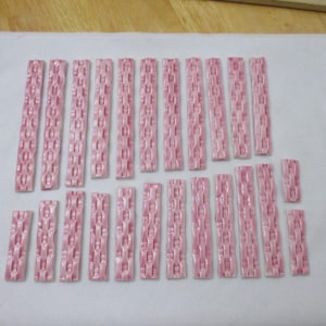 May include: A collection of pink ceramic tiles, each with a chain-link design, arranged on a white cloth. The tiles are rectangular and of varying lengths. The background is a light wooden surface.