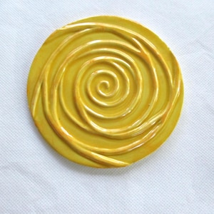 May include: A round, yellow ceramic coaster with a spiral design. The coaster has a raised, textured pattern in a lighter shade of yellow, creating a three-dimensional effect. The coaster is approximately 4 inches in diameter.