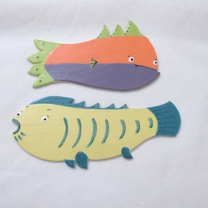 May include: Two whimsical, colorful fish decorations. One is orange and purple, the other is yellow and blue. Both have cartoonish features and are likely made of ceramic or painted wood. The fish are set against a white background.