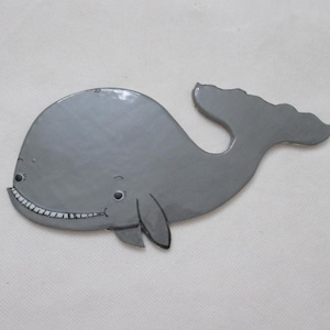 May include: A gray whale-shaped decorative item with a smiling face and a tail fin. The whale has small black eyes and fins. The design is simple and cartoonish, suitable for children's decor or nautical themes.