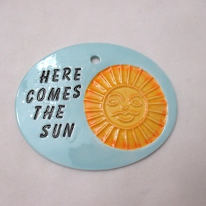 Puede incluir: Un óvalo de cerámica azul claro con el texto "HERE COMES THE SUN" en negro. Presenta un diseño de sol sonriente en amarillo y naranja. El óvalo tiene un agujero en la parte superior para colgar.