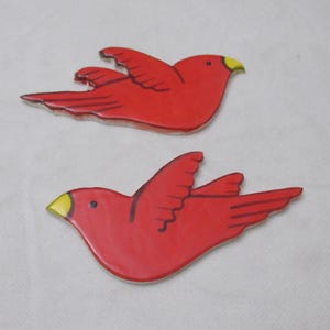 May include: Two ceramic bird figurines in flight. The birds are red with yellow beaks and black detailing. The wings are outstretched, and the birds are set against a white background.