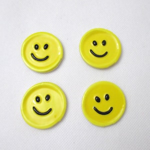 Puede incluir: Cuatro posavasos de cerámica amarillos con caras sonrientes. Cada posavasos presenta una base amarilla brillante con un diseño de cara sonriente negra. Los posavasos son redondos y tienen un borde ligeramente elevado.