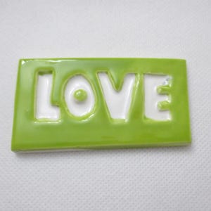 May include: A rectangular, lime green ceramic tile with the word "LOVE" in white, cut-out letters. The tile has a glossy finish and is set against a white background. The letters are bold and easy to read.