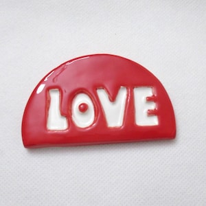 May include: A red, semi-circular decorative item with the word "LOVE" in white, bold letters. The item has a glossy finish and is set against a white background. The design is simple and eye-catching.