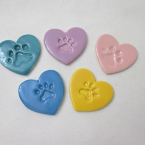 May include: Five heart-shaped ornaments in teal, lavender, light pink, blue, and yellow. Each smooth, glossy heart features a debossed paw print impression in the centre, creating a uniform set of decorative items with a consistent heart shape.