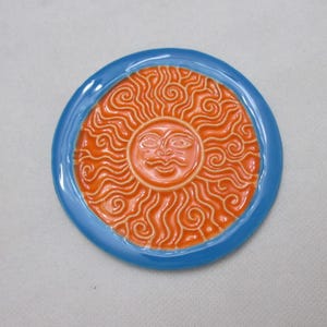 Op de afbeelding: Een keramische onderzetter met een fel oranje zonontwerp. De zon heeft een gezicht en is omringd door draaiende lijnen. De onderzetter heeft een lichtblauwe rand.