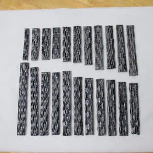 May include: A collection of dark grey metal rectangles, each with a textured chain pattern, are arranged on a white surface. The pieces vary in length and are positioned in different directions. The metal has a reflective quality.