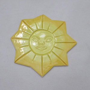 May include: A yellow ceramic sun-shaped ornament. The sun features a smiling face at its centre and pointed rays radiating outwards. The piece has a glossy finish and is likely a wall hanging or decorative tile.
