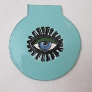 May include: A circular, light blue ceramic object with a flat top tab. The center features a stylized eye graphic with a blue iris, a green eyelid, and a black, sunburst-like border surrounding the eye, all finished with a glossy glaze.