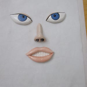 May include: A whimsical face composed of ceramic features. Two white-based eyes with blue irises and black outlines are positioned above a small nose and a smiling mouth with white teeth. The face is set against a white background.