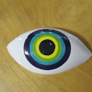 May include: White ceramic eye with a blue, green and yellow concentric circle design. The eye is painted in a traditional evil eye style.