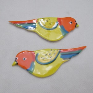 May include: Two ceramic bird figurines. Each bird has a yellow body, orange head, and red, blue, and yellow accents on the wings and tail. The birds are lying flat against a light gray background.