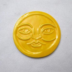 Puede incluir: Un rostro de sol redondo de cerámica amarilla con rasgos faciales en relieve. El sol tiene los ojos cerrados, una nariz y una boca sonriente. La pieza de cerámica tiene un acabado brillante y está centrada sobre un fondo blanco.