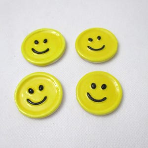 May include: Four yellow ceramic smiley face magnets. Each magnet has a round shape with a black smiling face. The magnets are arranged in a square pattern against a white background.