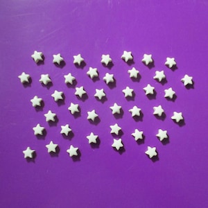 May include: White star-shaped sprinkles scattered on a vibrant purple surface. These small, decorative sprinkles are arranged in a loose pattern, creating a playful and festive visual. Ideal for decorating baked goods.