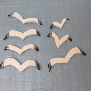 Può includere: Una collezione di sette piccoli ornamenti piatti a forma di uccello di varie dimensioni. Ogni pezzo è color bianco sporco con le punte delle ali nere, raffigurante un gabbiano stilizzato in volo con le ali angolate a forma di V.