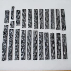 May include: A collection of small, rectangular, dark gray tiles with a textured, chain-like pattern. The tiles are arranged on a white surface, varying in size and quantity, suggesting a mosaic or decorative element.