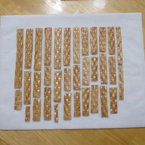 Puede incluir: Una colección de pequeñas piezas rectangulares de madera marrón claro con una textura tejida, dispuestas sobre un paño rectangular blanco. Las piezas de madera varían en tamaño y se colocan en un patrón similar a una cuadrícula. El fondo es una superficie de madera marrón claro.