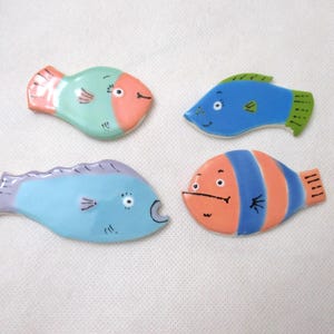 May include: Four ceramic fish-shaped ornaments featuring hand-painted details. One is mint green with a peach head, one is blue with a green fin, one is light blue with a purple tail, and one is orange with blue vertical stripes and black eyes.