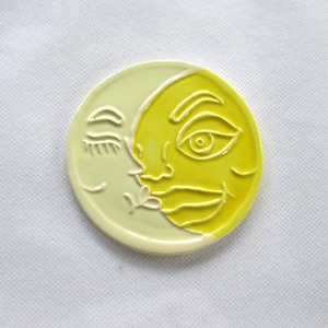 Può includere: Un pezzo di ceramica rotondo bicolore con un disegno di sole e luna. Il sole è giallo, con una faccia sorridente, e la luna è giallo pallido. Il disegno è in rilievo, creando un effetto strutturato. Probabilmente un oggetto decorativo.