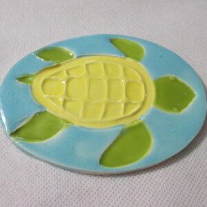 May include: A ceramic tile with a blue background and a yellow turtle design. The turtle has green flippers and a shell with a grid pattern.