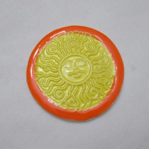 May include: A circular, yellow sun-shaped ornament with an orange rim. The sun features a smiling face and swirling rays. The item is likely ceramic or glass, and is a decorative piece.