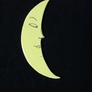 May include: A yellow crescent moon with a black outline and a face drawn on it. The moon is a ceramic wall hanging.