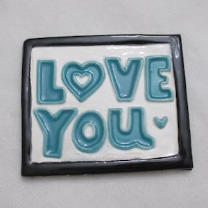 Puede incluir: Una baldosa de cerámica con las palabras "LOVE YOU" en letras turquesas sobre un fondo blanco, enmarcada por un borde negro. El corazón en "LOVE" también es turquesa.