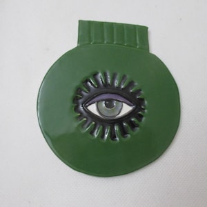 Puede incluir: Un adorno de pared de cerámica verde de forma circular con un motivo de ojo central. El ojo tiene un iris gris claro, una pupila negra y un párpado morado, rodeado por un patrón de rayos de sol negro. Una pestaña verde rectangular sobresale arriba.