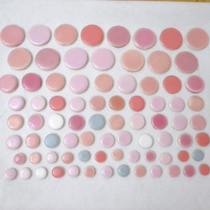 May include: A collection of numerous small, round, flat ceramic tiles arranged in rows. The tiles feature a glossy finish and a color palette consisting of various shades of pink, peach, coral, white, and light blue, organized by size.