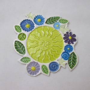 May include: A ceramic wall plaque featuring a central lime green sun face with wavy rays, surrounded by a white border decorated with blue, purple, and light blue flowers, green leaves, and a small green peace sign symbol.