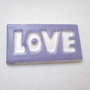 May include: A rectangular ceramic tile with the word "LOVE" in white, set against a lavender background. The letters are cut out, creating a positive and negative space effect. The tile is smooth and glossy.