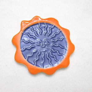 May include: A circular ceramic dish featuring a central sun face with wavy rays in a glossy periwinkle blue finish. The outer border is a scalloped, vibrant orange rim. The piece has a smooth, glazed texture and a distinct relief design.