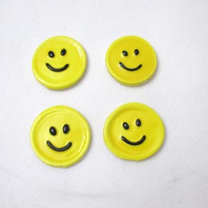 Könnte beinhalten: Vier gelbe Keramik-Untersetzer mit Smiley-Gesichtern, schwarzen Augen und einem gebogenen Mund. Die runden Untersetzer haben einen leicht erhöhten Rand. Der Hintergrund ist eine weiße, strukturierte Oberfläche.