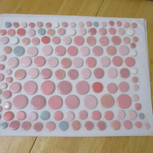 May include: A collection of pink, white, and gray buttons of varying sizes arranged on a white background. The buttons range in shades from light pink to coral, with a few gray and white accents. The buttons are all circular.