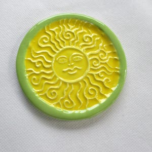 May include: A round ceramic dish featuring a bright yellow center with a raised, embossed sun face design. The sun has wavy, swirling rays extending to the edge. The dish is framed by a smooth, solid light green border, creating a distinct contrast.