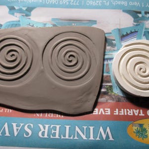 Könnte beinhalten: Ein grauer Tonstempel mit zwei Spiralmustern und ein separater weißer Stempel mit einem einzelnen Spiraldesign. Der Ton befindet sich auf einer bedruckten Oberfläche mit dem Text "WINTER SAVING".