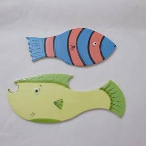May include: Two ceramic fish decorations. One is blue with pink stripes and black accents. The other is yellow with green fins and details. Both have whimsical, cartoon-like features and are set against a white background.