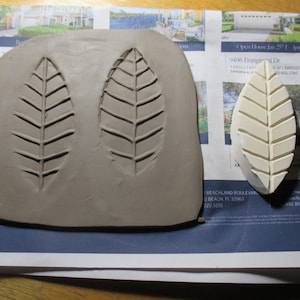 May include: A clay leaf stamp and a clay impression of two leaves. The stamp is off-white and leaf-shaped, with detailed veining. The clay impression is a light brown color, showing the negative space of the leaf design.