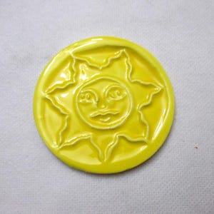 Op de afbeelding: Een ronde, gele keramische onderzetter met een lachend zonontwerp. De zon heeft een gezicht met ogen, een snor en stralende stralen. De onderzetter is glanzend en heeft een verhoogd ontwerp.