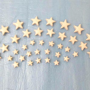 Könnte beinhalten: Anordnung cremefarbener sternförmiger Dekorationen in verschiedenen Größen. Die Sterne sind auf einer hellblauen Oberfläche verteilt und erzeugen eine himmlische oder skurrile Ästhetik. Die Sterne sind wahrscheinlich aus Holz oder einem ähnlichen Material.