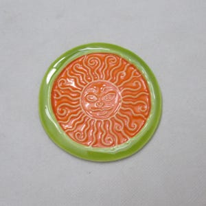 May include: A ceramic sun with a green rim and an orange face. The sun has a stylised face with a smile and swirls for hair.