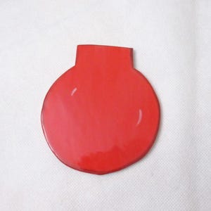 Puede incluir: Adorno decorativo rojo brillante, con cuerpo redondeado y cuello corto y rectangular. El adorno tiene una superficie lisa y reflectante, sobre fondo blanco. La forma recuerda a un jarrón o botella estilizada.